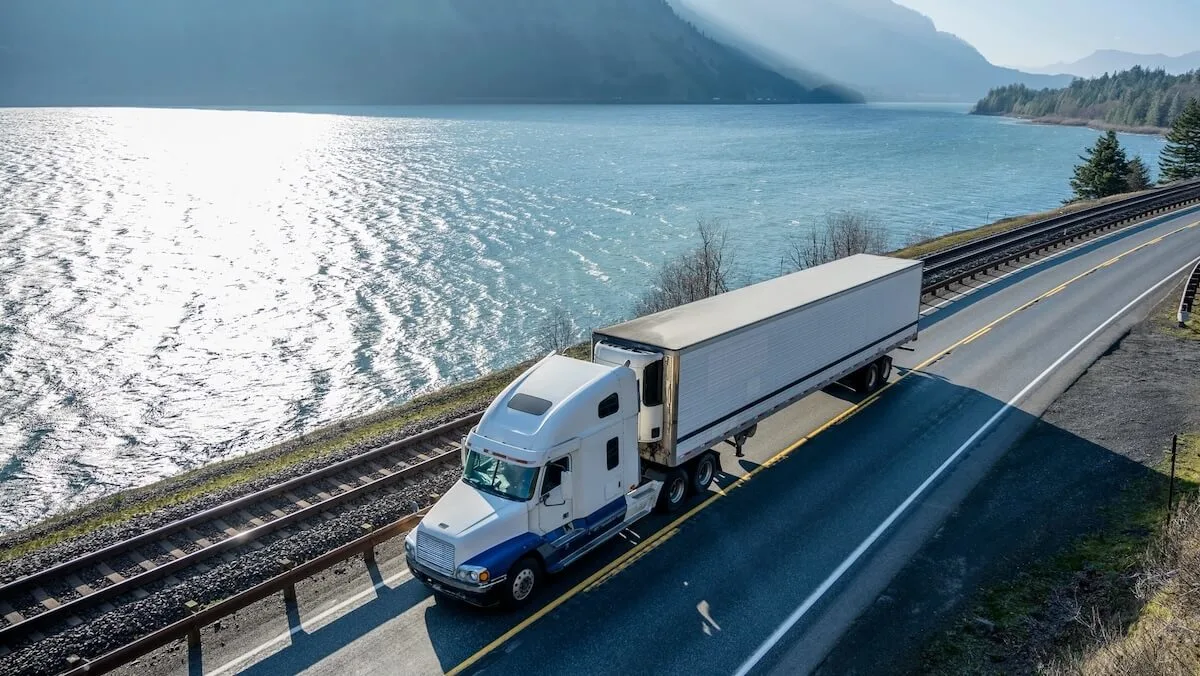 White semi-truck hauling a trailer on a lakeside highway with mountains in the hazy distance.