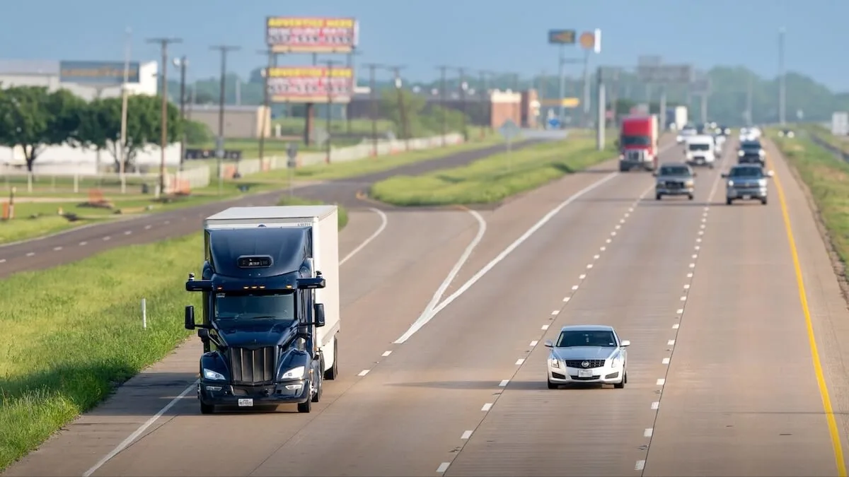 A high-angle, front-facing view of a dark blue semi-truck with a white trailer driving on a multi-lane highway alongside a silver sedan, with other distant traffic and billboards visible under a clear sky.