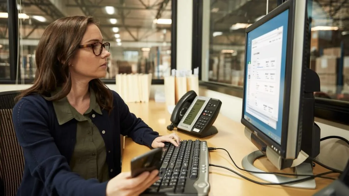 Logistics office worker using a desktop computer and holding a phone while reviewing shipment information on screen at a warehouse facility.
