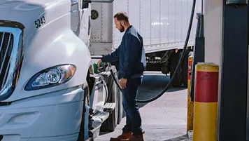 Truck driver refueling a white semi-truck at a gas station, standing between the truck and fuel pump.