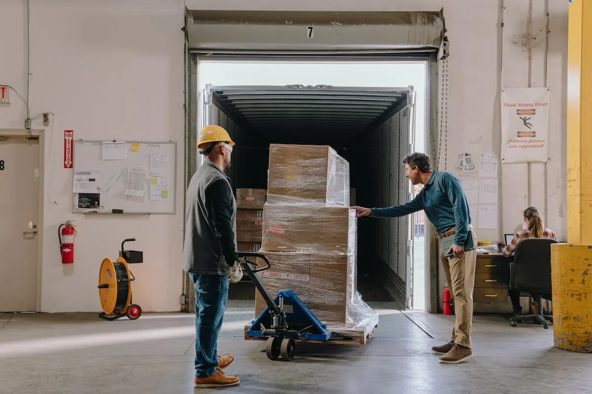 Deux employés d'entrepôt inspectent une palette de cartons emballée sous film plastique en cours de déchargement d'une remorque de camion à un quai de chargement.