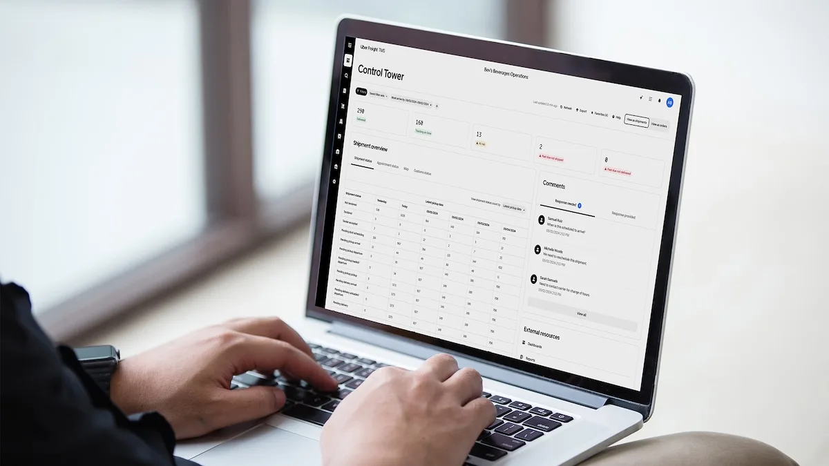 A person typing on a laptop displaying the Uber Freight TMS Control Tower dashboard, showing real-time shipment status metrics, network tracking data, and communication logs.