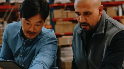 Two warehouse workers reviewing documents and plans together in a storage facility with shelves in background.