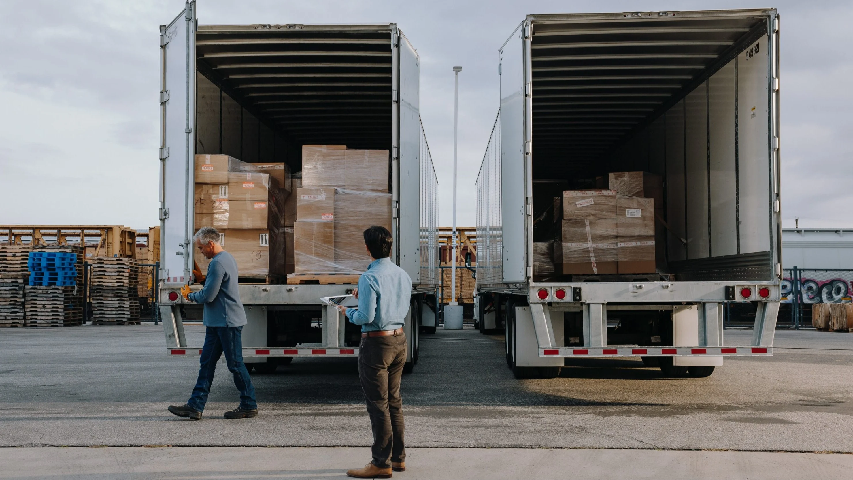 Two workers at a shipping yard with open semi-trailers loaded with boxed cargo and wooden pallets nearby.