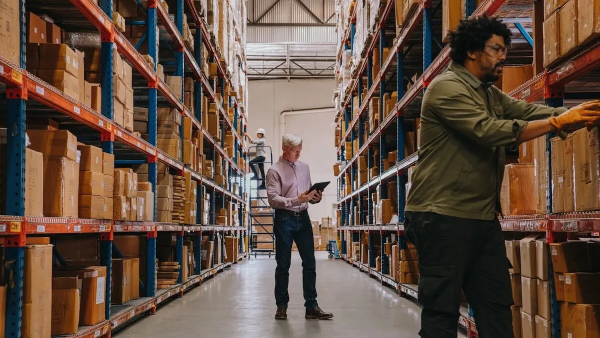 Trois employés dans une allée d'un grand entrepôt inspectent et organisent des boîtes en carton sur de hautes étagères à palettes, deux d'entre eux utilisant des tablettes tandis que le troisième atteint des marchandises sur l'étagère.
