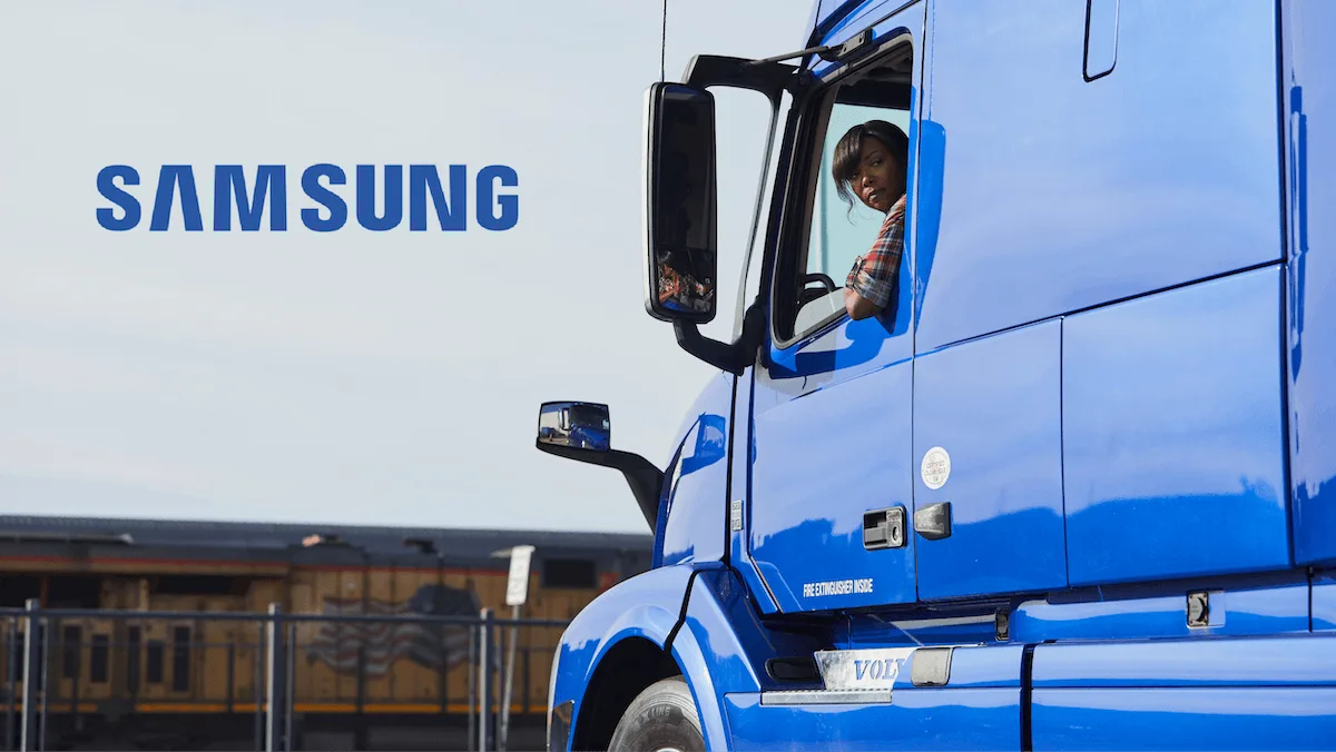 Truck driver looking out the window of a bright blue semi‑truck cab with a freight train in the background.