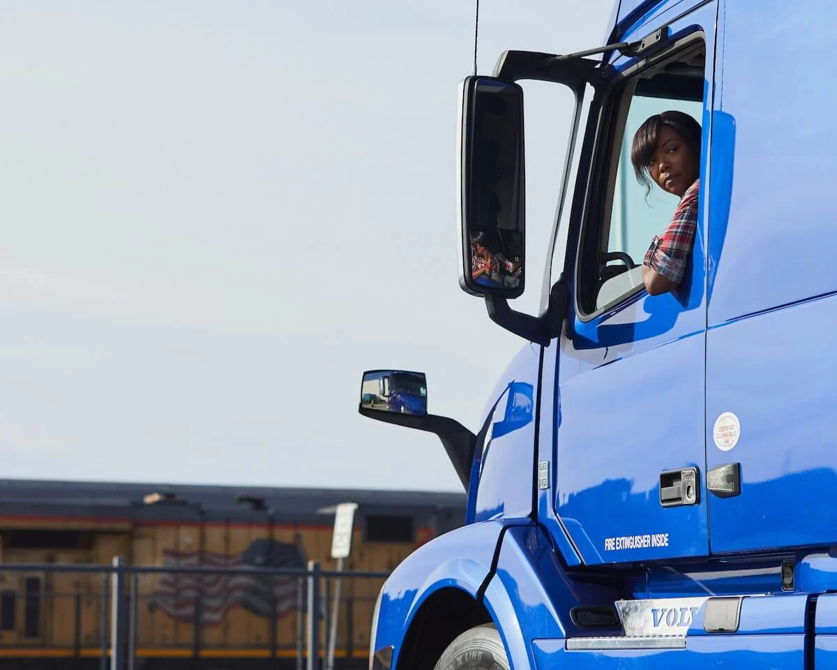 Truck driver looking out the window of a bright blue semi‑truck cab with a freight train in the background.