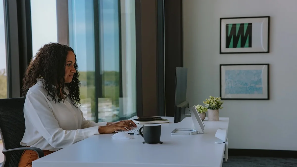 Woman sitting at a modern office desk working on a desktop computer near large windows, with framed artwork on the wall and a plant and coffee mug.