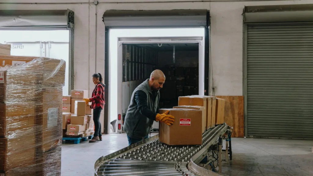Workers in warehouse handling cardboard boxes on conveyor belt with pallets and loading dock in background.
