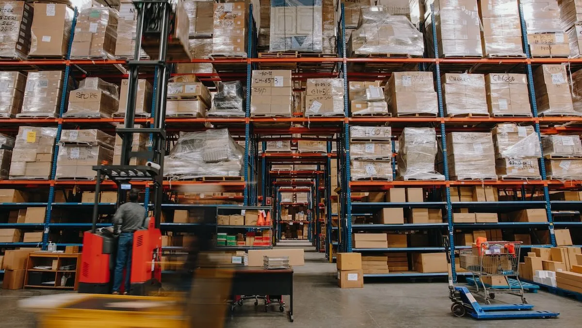 Busy warehouse interior with tall pallet racks stacked with boxes, a worker operating a red forklift, and blurred motion from another vehicle in the foreground.