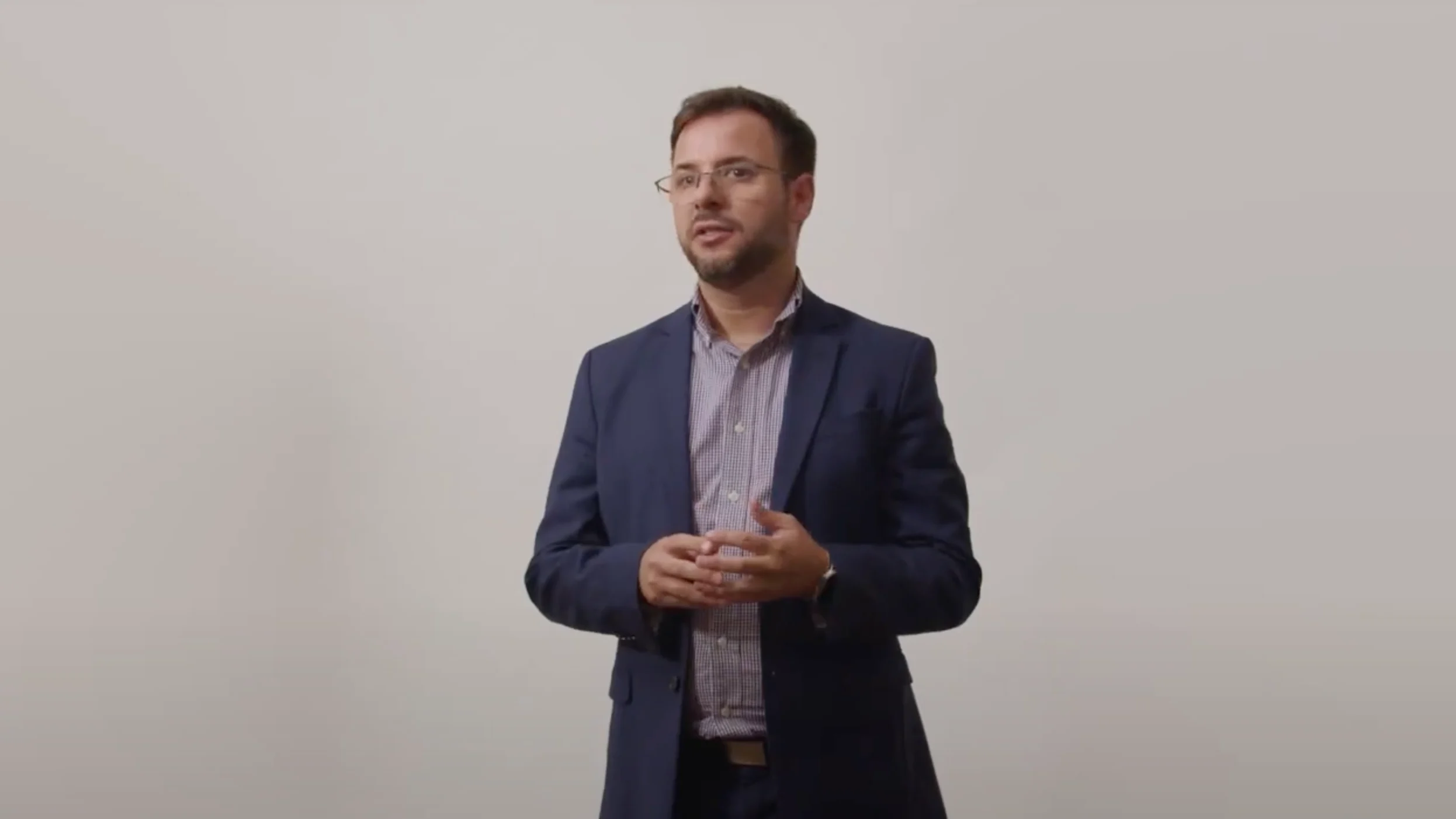 Video thumbnail with play button overlay on a person in a navy suit against a light gray background.