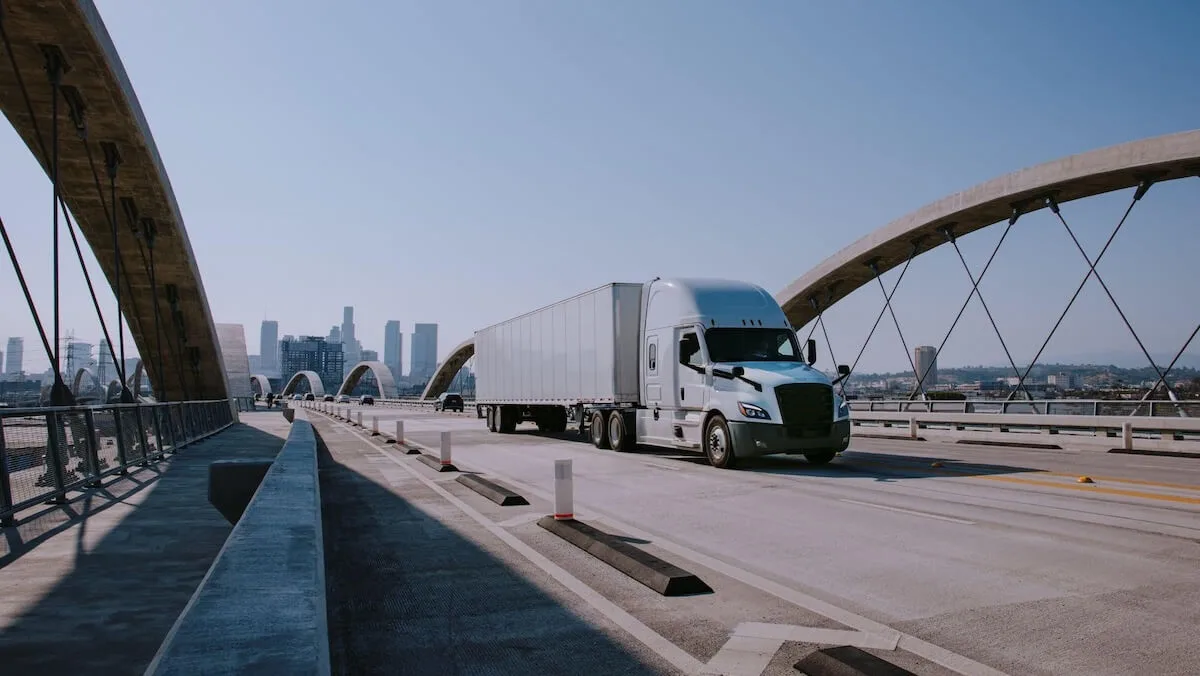White truck driving over a bridge with a city in the background