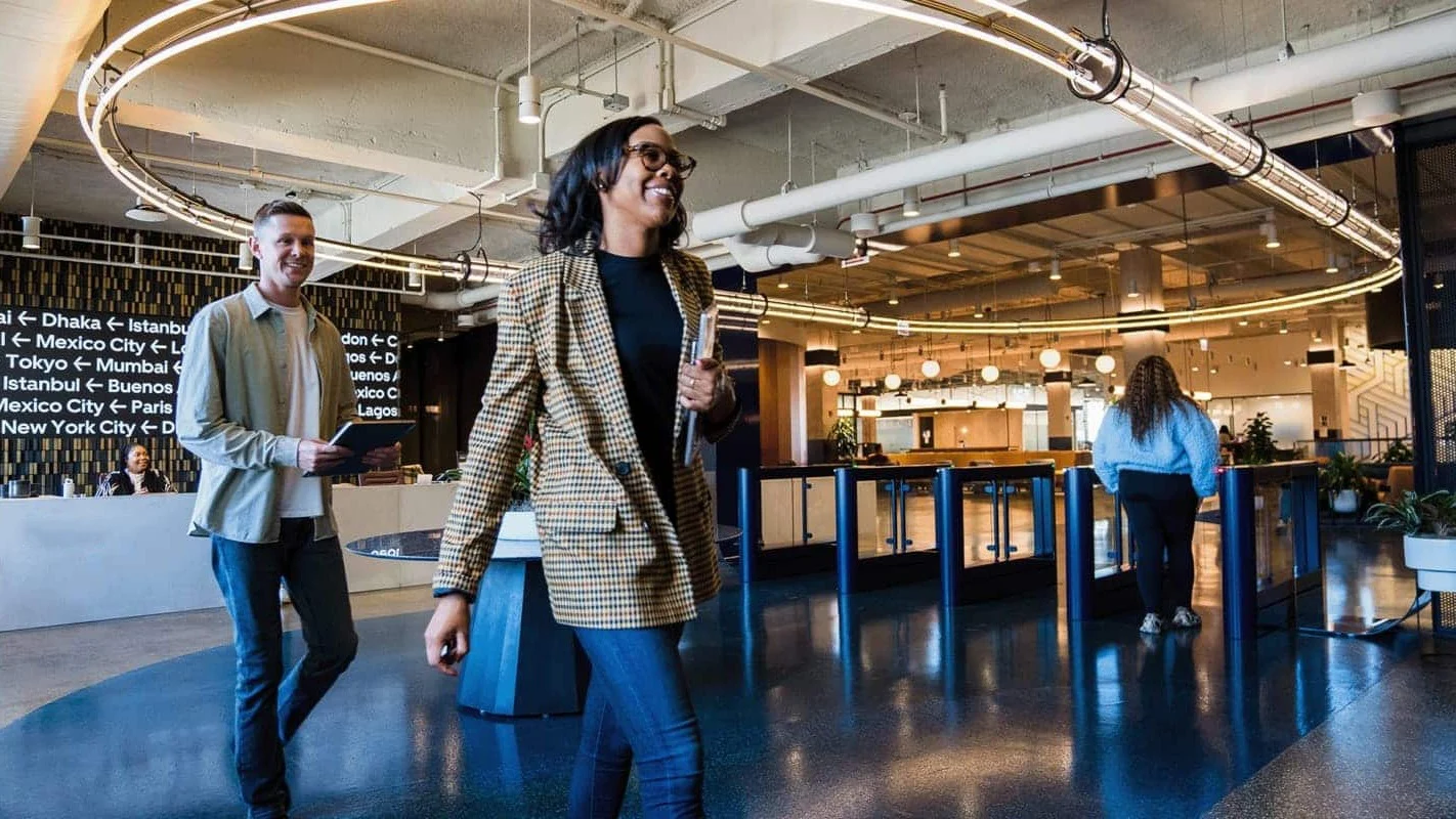 People walking through modern office space with circular lighting fixture, global city names on wall, and reception area.