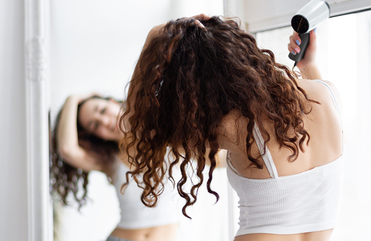 Young brunette girl female woman adult in grey shorts and white top drying her curly hair with blow dryer after shower in front of the mirror.