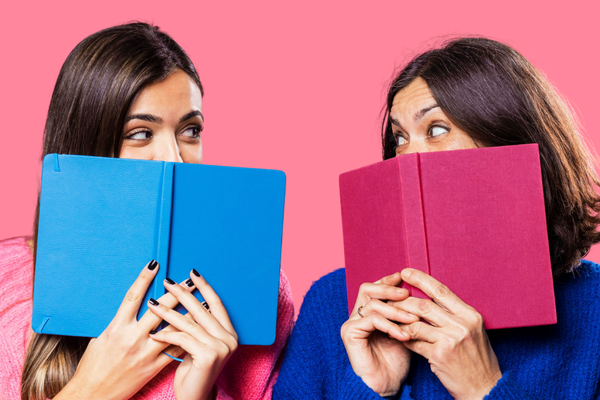 Two smiling women hold colorful books—one blue, one red—while making eye contact over them against a bright pink background.