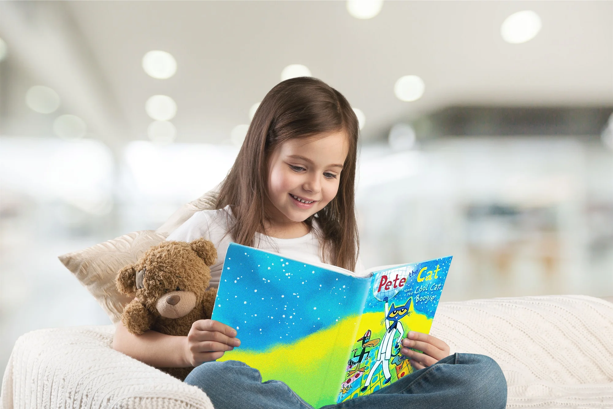 Photo of little girl holding a teddy bear, reading a Pete the Cat book while sitting on a couch.