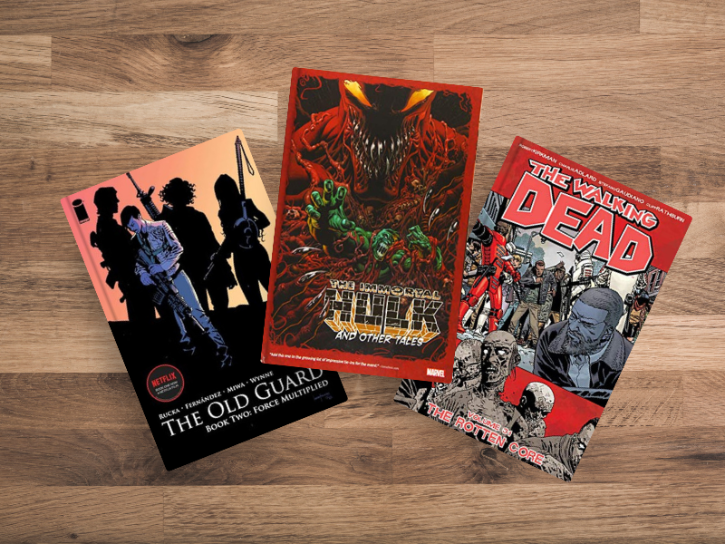 "Three comics & graphic novels on wooden surface: The Old Guard by Greg Rucka (black cover with silhouetted warriors), The Immortal Hulk and Other Tales (red cover with menacing Hulk creature), and The Walking Dead by Robert Kirkman (red cover with zombie apocalypse scenes)."