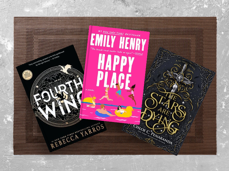Three Bestsellers of 2025 So Far books arranged on woven mat: Fourth Wing by Rebecca Yarros (black cover with circular design), Happy Place by Emily Henry (bright pink cover with beach scene), and The Stars Are Dying by Chloe C. Peñaranda (dark ornate cover with golden decorative elements).