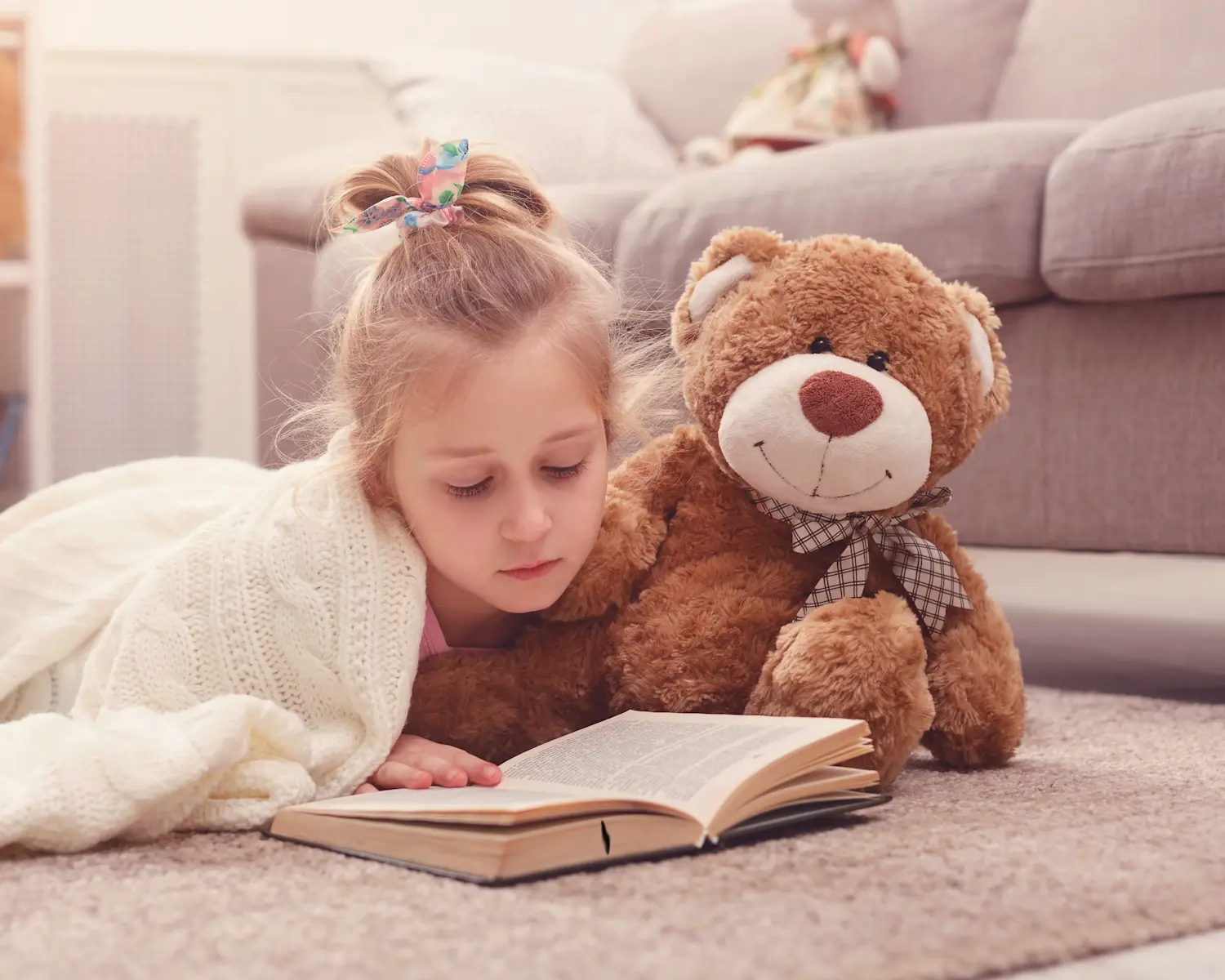 Cozy reading time with child enjoying book on carpeted floor accompanied by brown teddy bear in comfortable living space