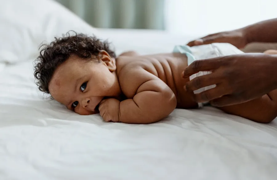 Baby laying on a bed with a parent's hands reaching in to pick the baby up.
