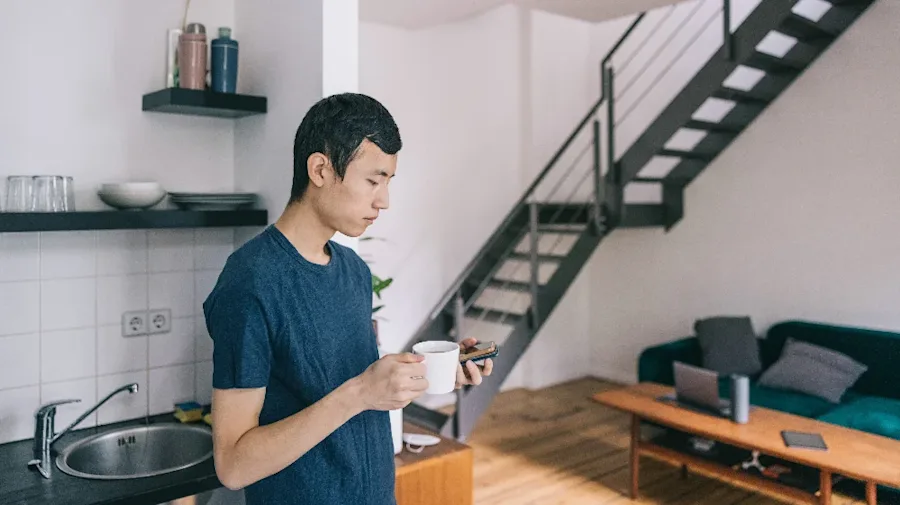 A person holding a coffee cup and their phone in an apartment.