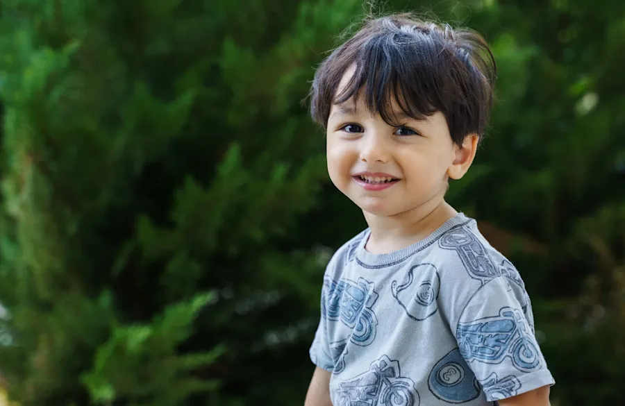 A young boy smiling outside in front of greenery.