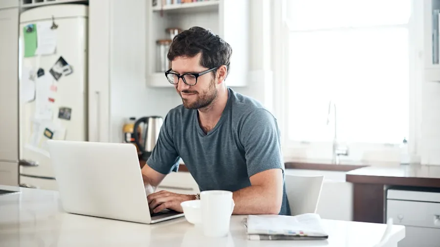 A man seated at home, smiling while interacting with a laptop.