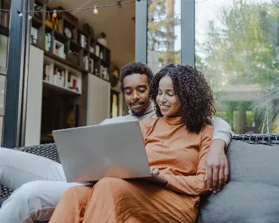 A couple seated on a couch, smiling at a laptop.