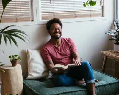A man seated on a couch smiling, reading a book.