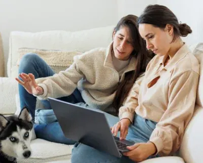 Two females seated on a couch looking at a laptop. One female is reaching to pet a cute dog.
