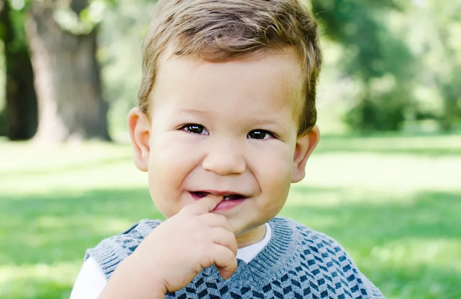 A toddler seated outside, smiling, with a finger in his mouth.