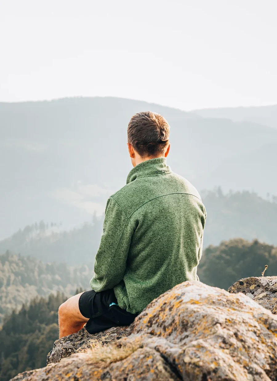 A mean seated atop a mountain looking at trees below.