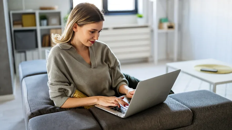A person smiling, seated on a couch interacting with a laptop.