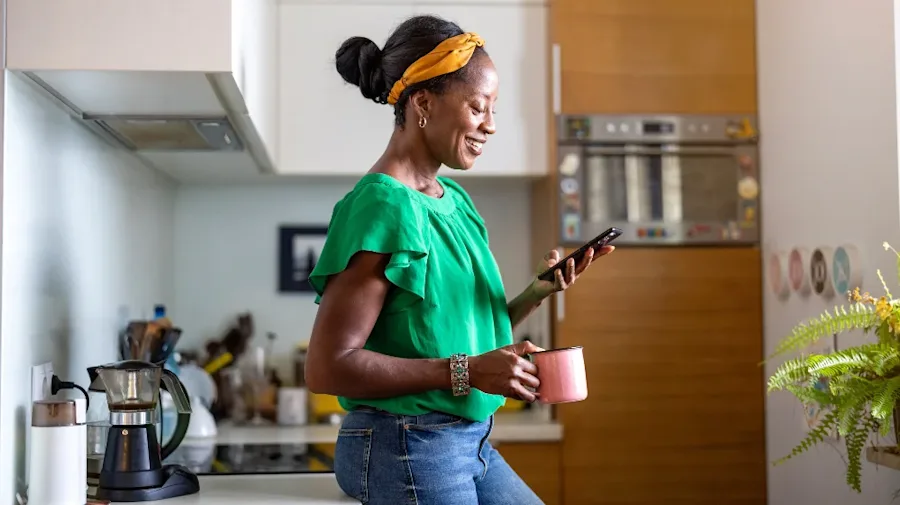 A woman holding a mug, smiling while looking at their phone.
