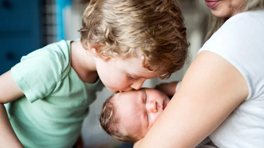 Person holding an infant while a child kisses the infant's forehead.