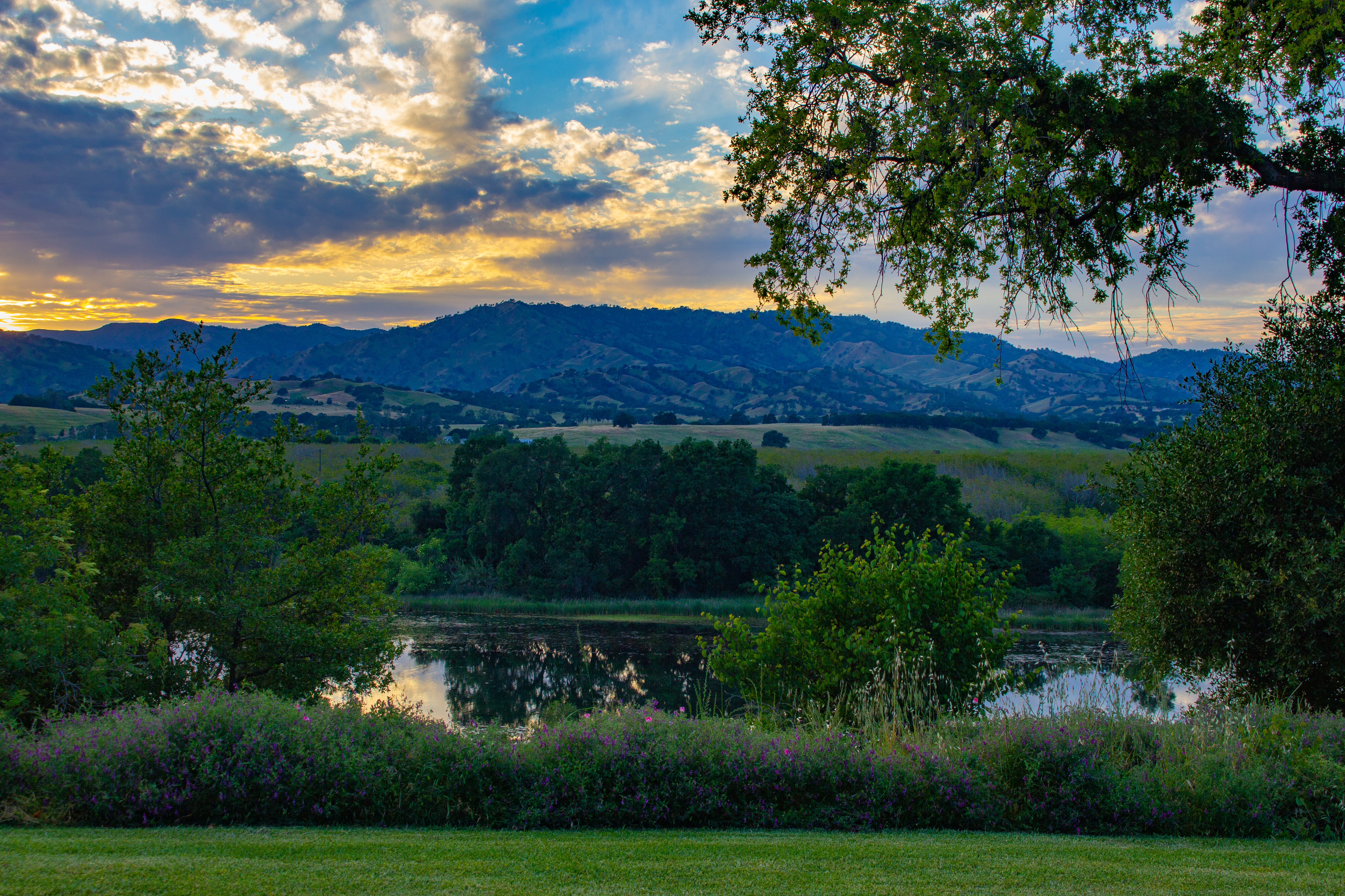 A photograph of clouds above a mountain, beyond a river, during sunset.