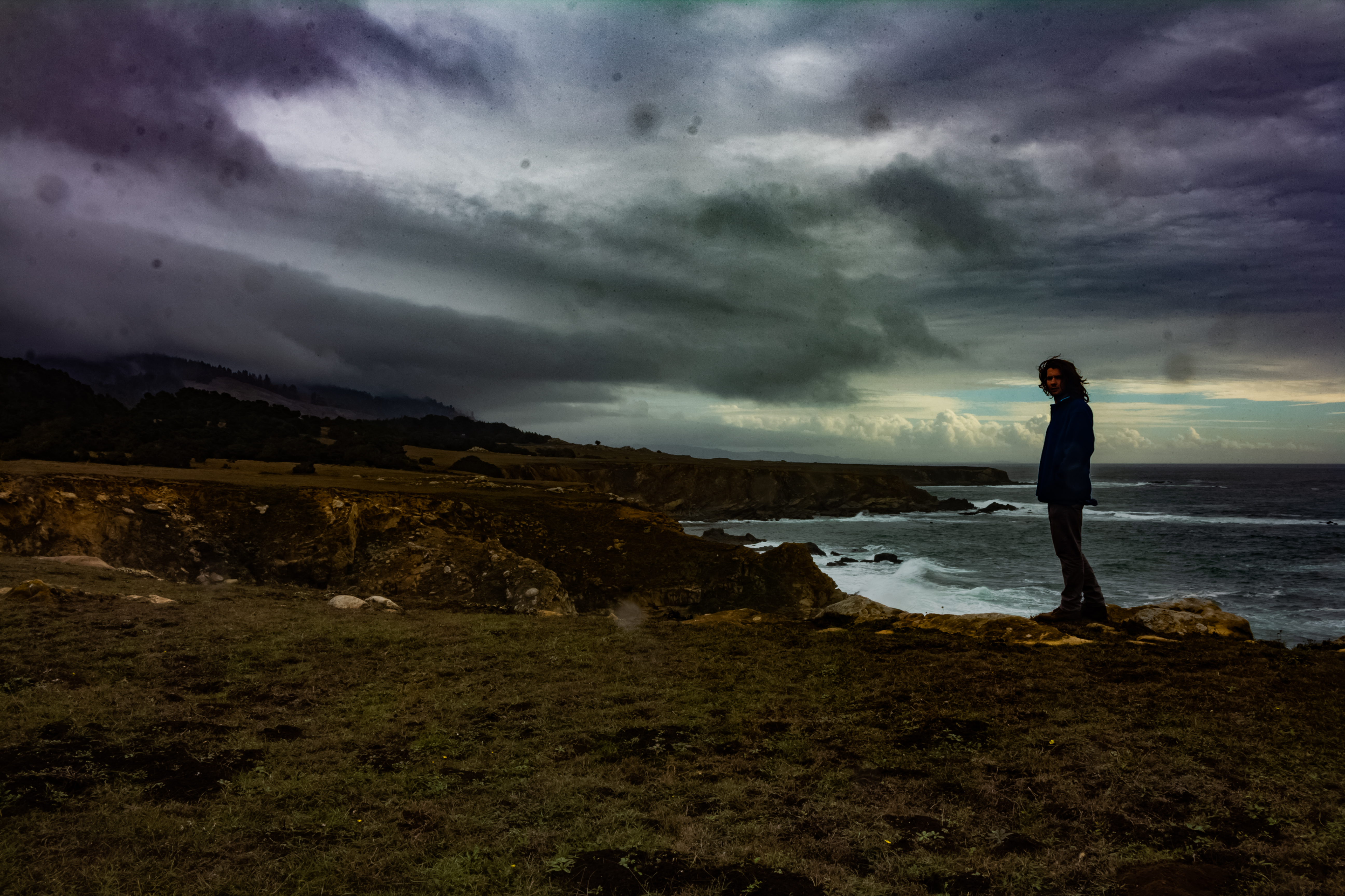 A photograph of the coast, with a person standing on the edge of a cliff looking back at the camera.