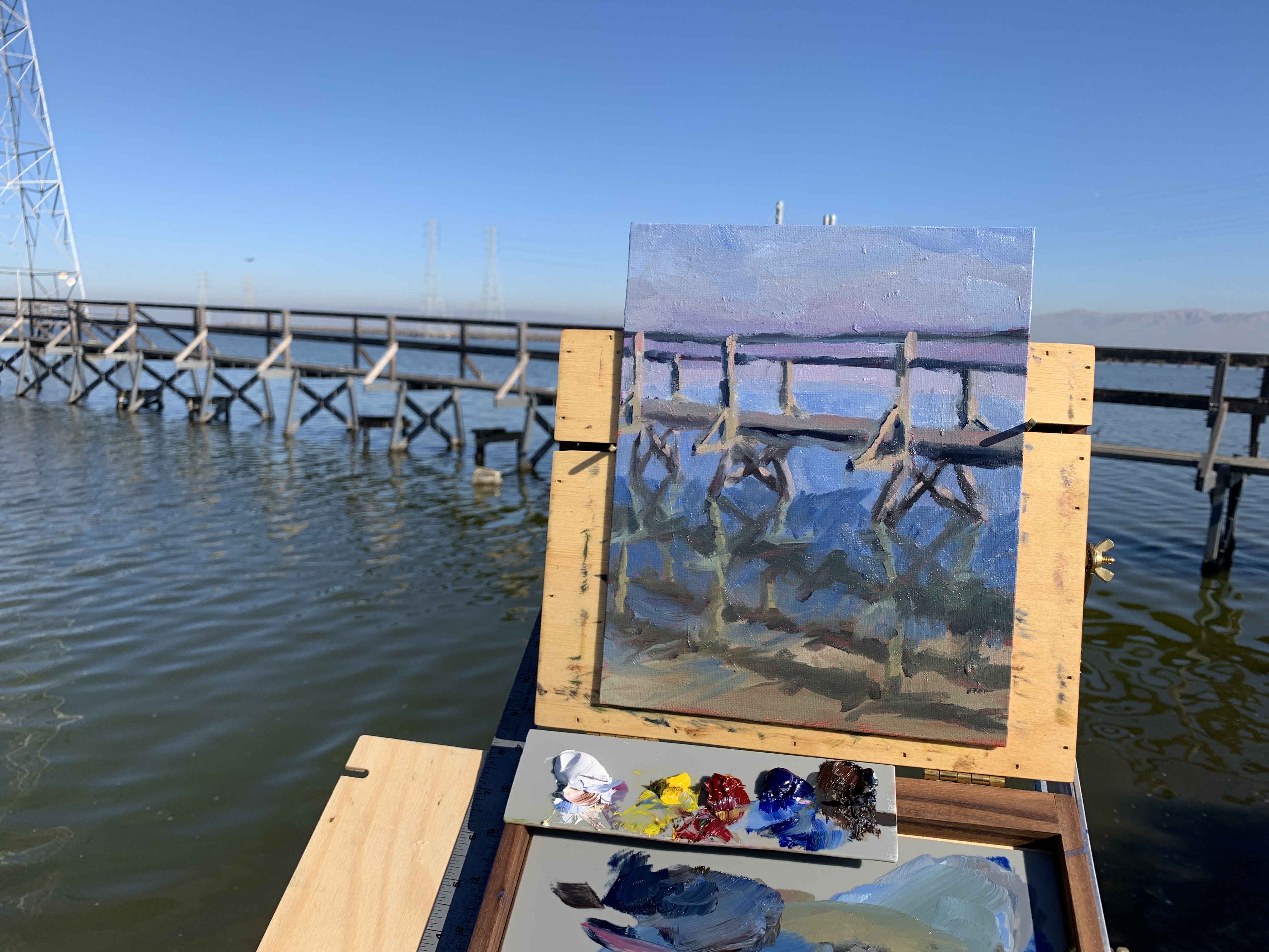 A photograph of a painting in the context of the scene that it depicts: A wooden bridge over some water on a sunny day.