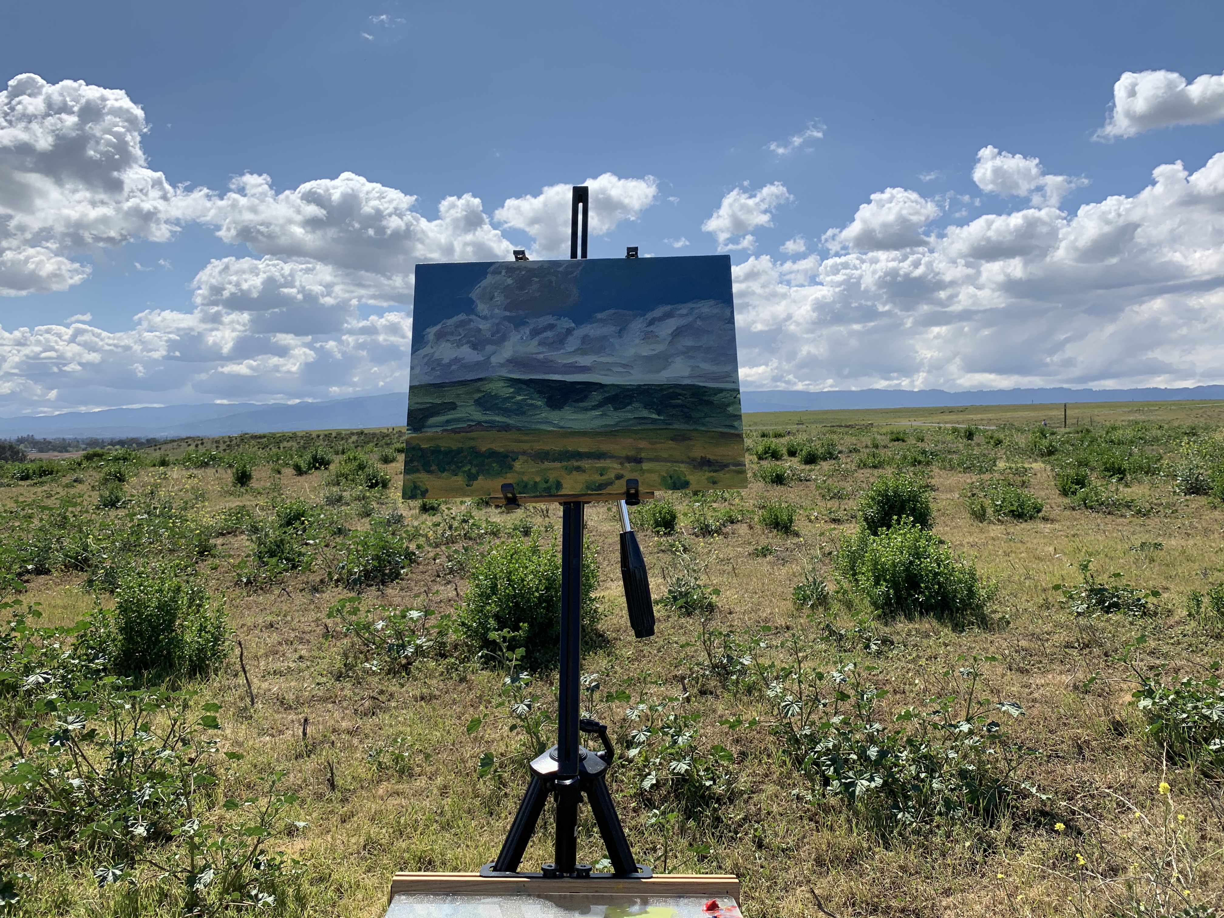 A photograph of a painting shown in the context of the scene it depicts: A distant hill shadowed by clouds on a sunny day.