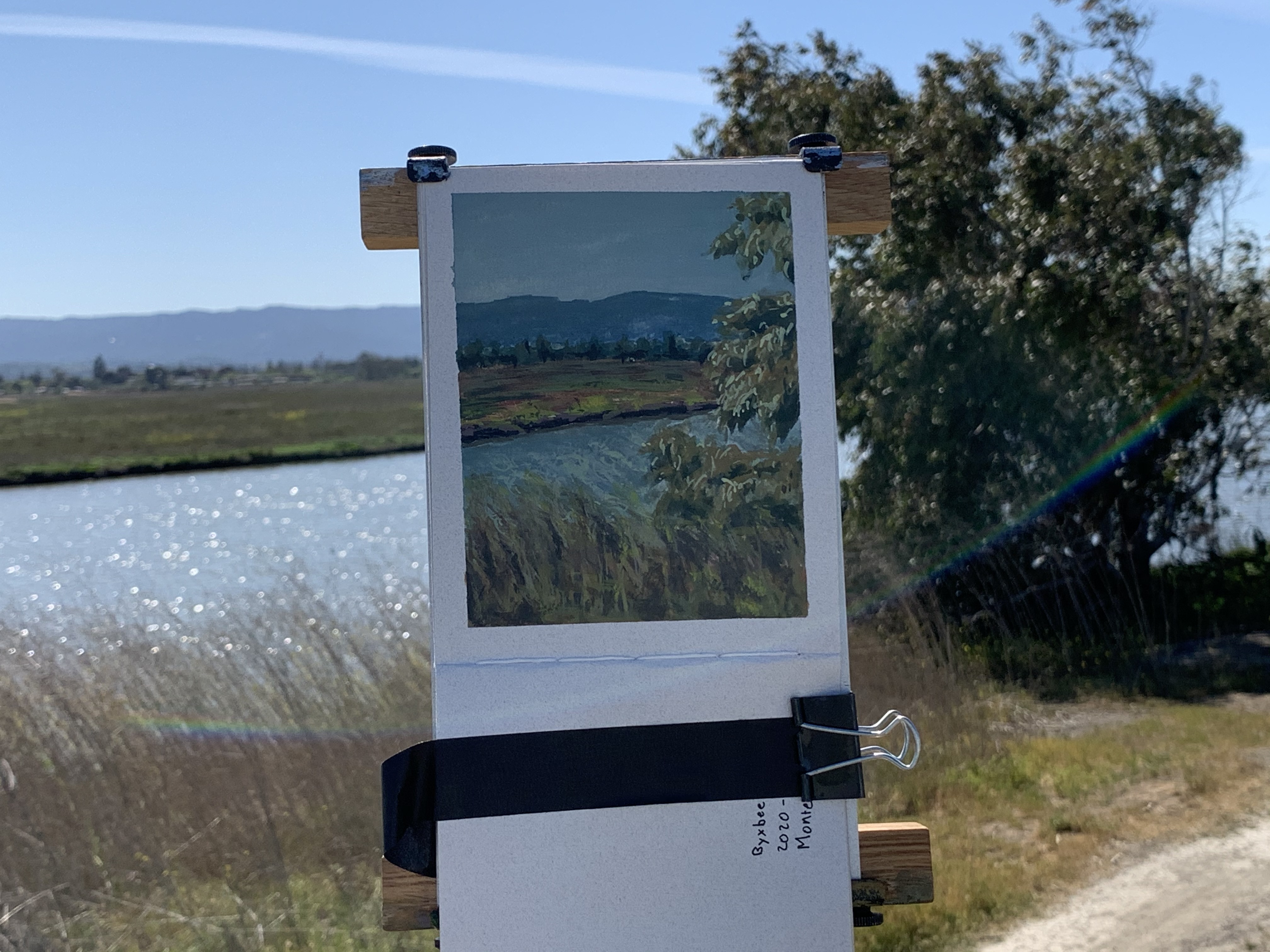 A photograph of a painting shown in the context of the scene it depicts: A river with a field and mountain beyond on a sunny day.