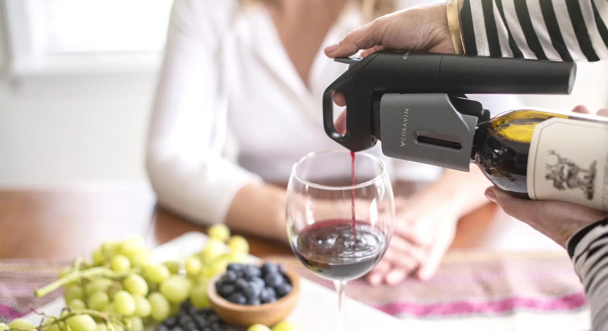 A woman pouring wine into a glass using the Coravin Model Three System with grapes, blueberries, and another woman in the background.