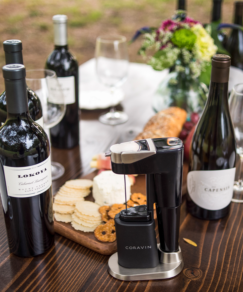 Plusieurs bouteilles de vin et d’encas disposés sur une table en extérieur, avec un système Coravin Model Eleven dans son socle.