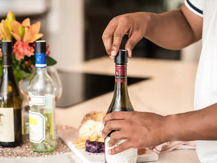 Person an einem Tisch, die einen Coravin Schraubverschluss auf eine Weinflasche schraubt, mit Fingerfood im Hintergrund

