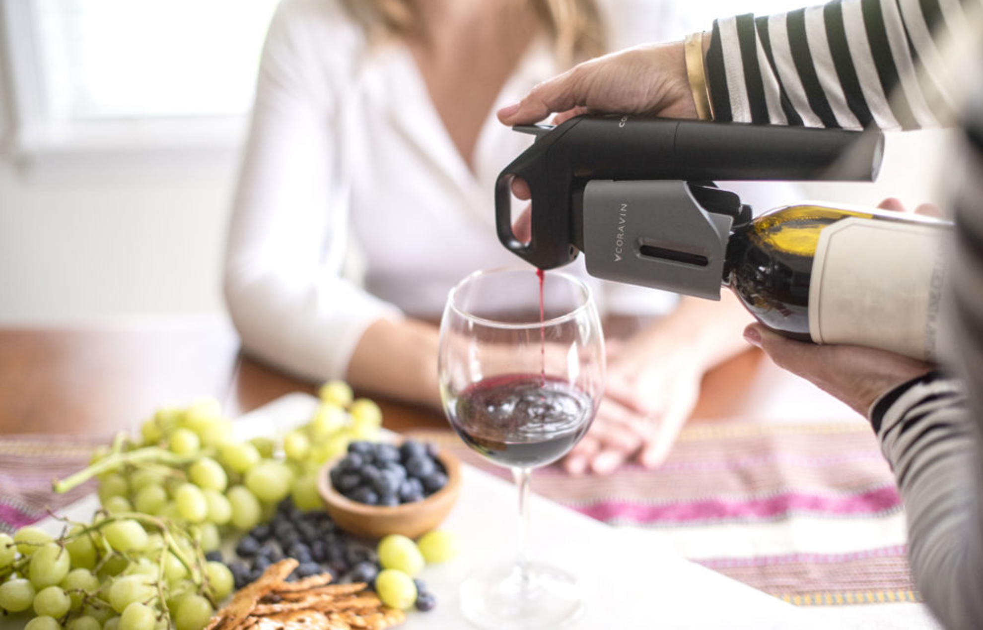 Vrouw die aan tafel zit waarop fruit ligt en een glas rode wijn staat, die wordt ingeschonken met een Coravin-wijnbewaarsysteem.
