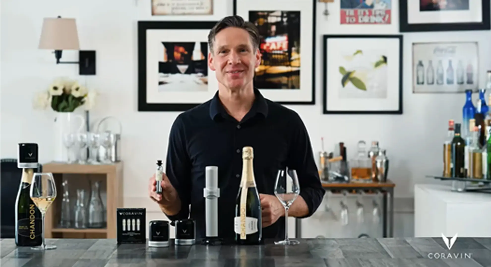 A man standing at a countertop holding a Coravin Pure CO2 Capsule in his hand. The Coravin Sparkling Preservation System is on the table in front of him, along with a bottle of wine. In the background are wine glasses, a bar cart, wine and liquor bottles, and multiple framed images.
