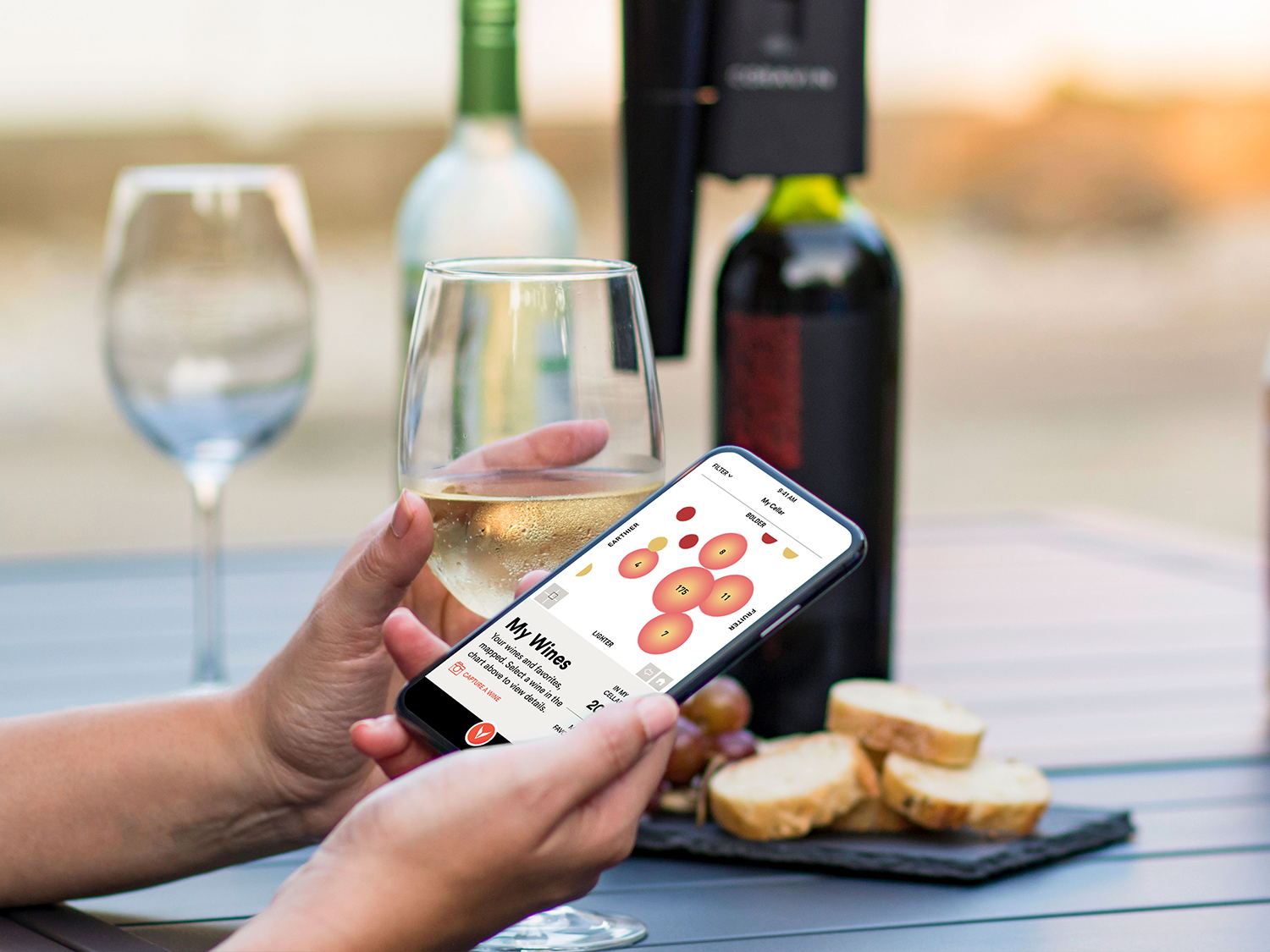Une personne tenant un verre de vin dans une main et un téléphone dans l’autre sur lequel s’affiche la carte des saveurs de l’application Coravin Moments, avec en arrière-plan le système de préservation du vin Coravin Model Eleven placé sur une bouteille.