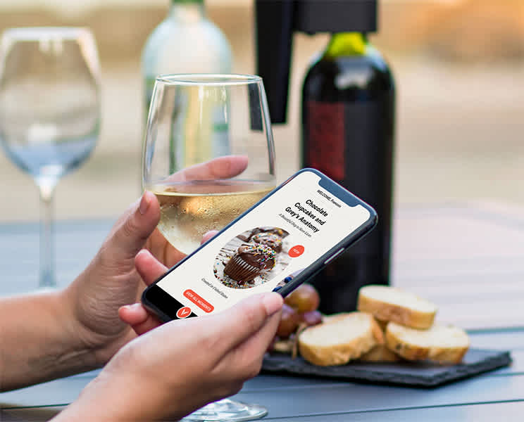Een persoon die een glas wijn vasthoudt en een telefoon met het scherm van de Coravin Moments-app met de smakenkaart, met op de achtergrond het Coravin Model Eleven-wijnbewaarsysteem op een fles.
