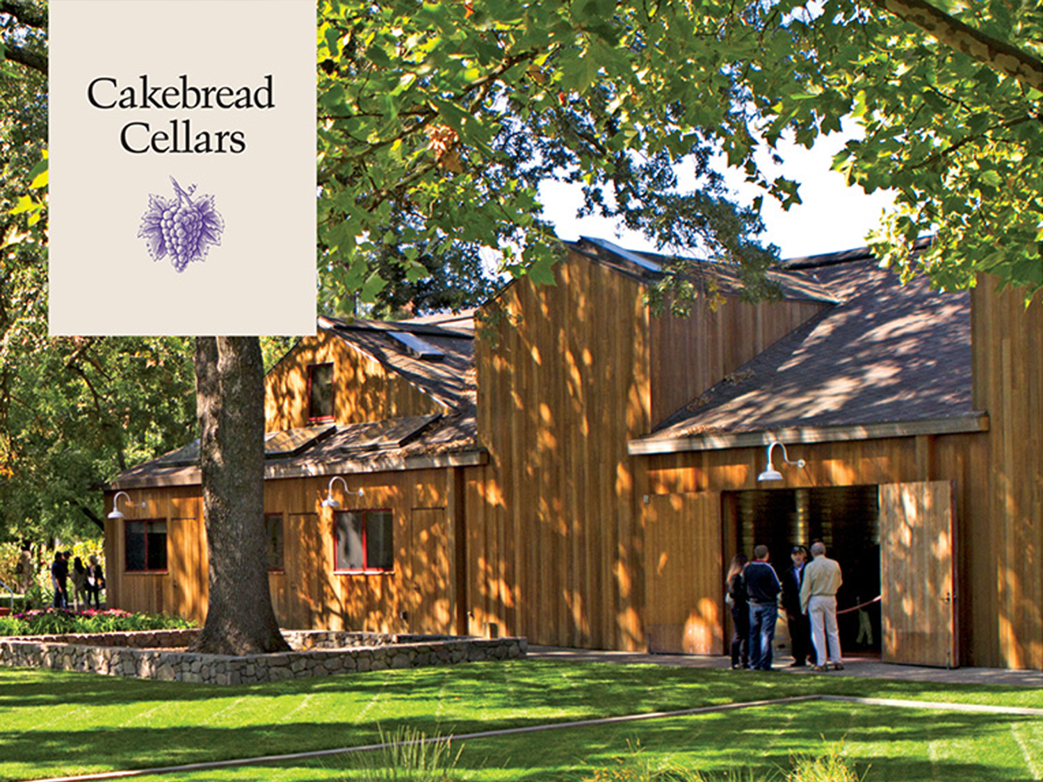 Image of a barn with green grass and people standing outside of it featuring the Cakebread Cellars logo.