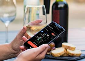 Een persoon die een glas wijn vasthoudt en een telefoon met het scherm van de Coravin Moments-app met Model Eleven, met op de achtergrond het Coravin Model Eleven-wijnbewaarsysteem op een fles.