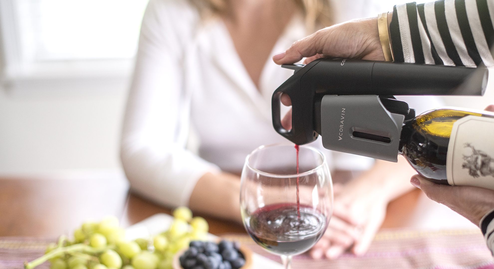 Woman sitting at a table spread of fruit with red wine being poured by a Coravin Wine Preservation System.